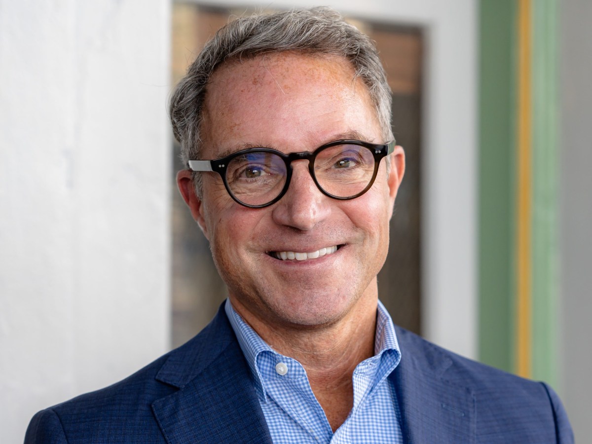 Hal Cato, wearing round glasses, a blue checked shirt and dark blue suit jacket, smiles as he stands in front of a gray door.